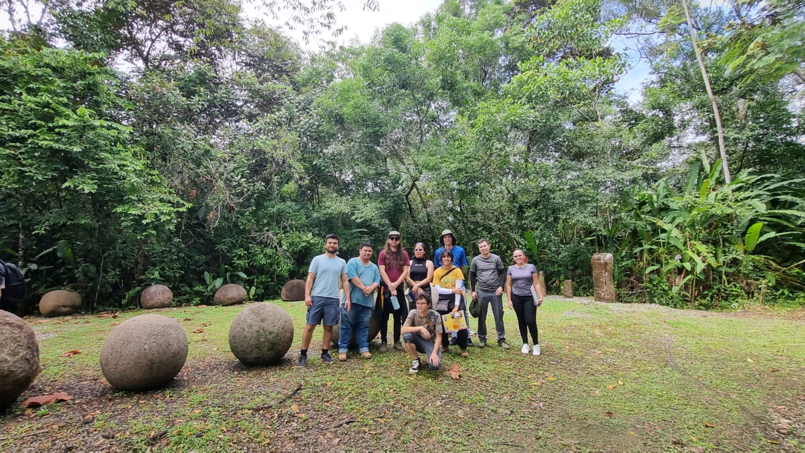 Nueve estudiantes posan para la foto al lado de una fila de esferas de piedra en Finca 6. Atrás, …