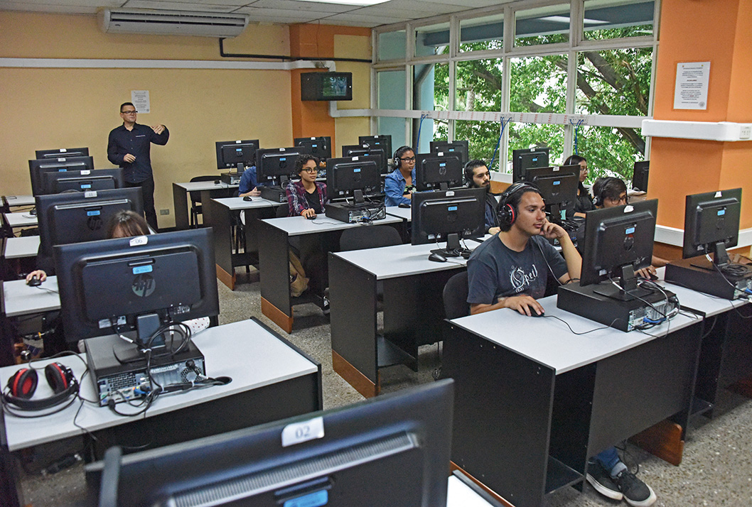 Un grupo de estudiantes aplican un examen en un laboratorio de cómputo. Atrás se aprecia al …