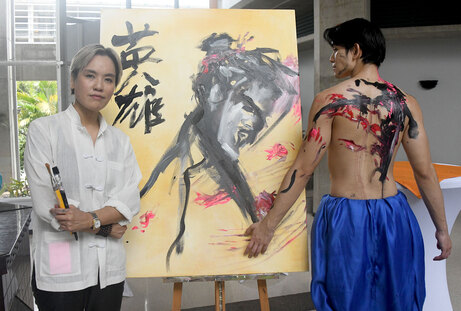 Una pintora con los pinceles en la mano posa al lado de su obra colocada en un caballete. Al otro …